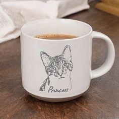 a white coffee mug with a cat drawn on the front and side, sitting on a wooden table