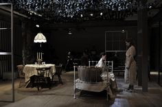 two women standing in a dark room next to a bed and table with white cloths on it