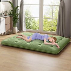 a woman laying on top of a green mattress in a room with windows and curtains