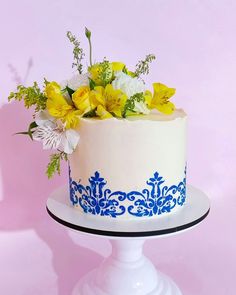 a white cake with blue and yellow flowers on top is sitting on a pedestal against a pink background