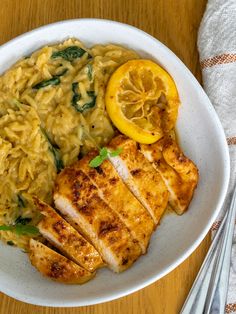 a white plate topped with chicken, pasta and lemon wedges on top of a wooden table