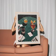 a woman sitting on a couch holding up a framed photo with two glasses and playing cards