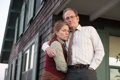 an older man and woman standing next to each other in front of a house with their arms around each other