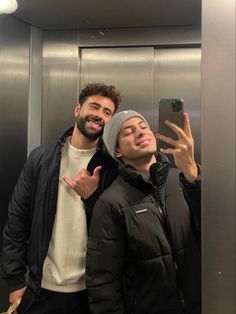 two men standing in front of an elevator taking a selfie with their cell phone