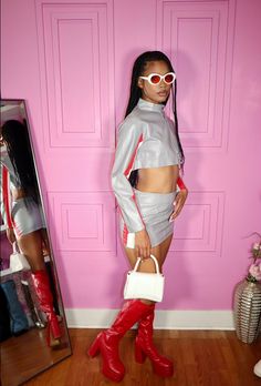 a woman standing in front of a pink wall wearing red boots and a silver top