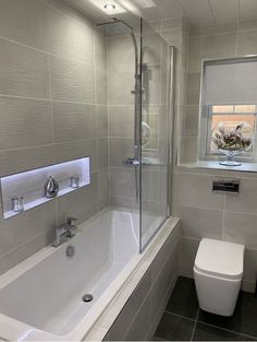 a white toilet sitting next to a bath tub under a window in a bathroom with gray tiles