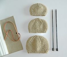 three knitted hats and two knitting needles on a white surface next to a notebook