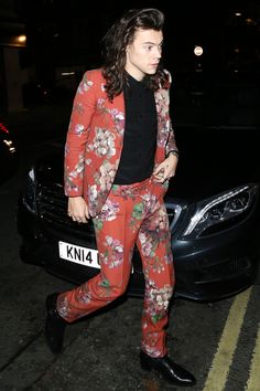 a woman in floral pants and a red blazer is standing next to a car