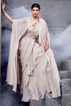 Ivory cape with floral embroidery and button detailing. Paired with pre-draped ruffle saree, embroidered blouse and embroidered belt.
Components: 4
Pattern: Embroidered
Type Of Work: Floral
Neckline: Round
Sleeve Type: Cape sleeves
Fabric: Georgette, net
Color: Ivory
Other Details: 
Embroidered belt detailing
Tassel detailing
Pleated detailing
Occasion: Wedding - Aza Fashions Luxury Pre-draped Saree With Cape Sleeves For Women, Traditional Pre-draped Saree With Cape Sleeves For Wedding, Elegant Lehenga With Draped Cape Sleeves, Festive Pre-draped Saree With Cape Sleeves For Wedding, Georgette Saree With Cape Sleeves For Reception, Georgette Saree With Resham Embroidery And Cape Sleeves, Elegant Pre-draped Saree With Cape Sleeves In Georgette, Elegant Ruffled Gown For Designer Wear, Traditional Lehenga With Draped Cape Sleeves