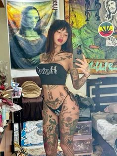 a woman with tattoos on her body is standing in front of a bed and holding a cell phone