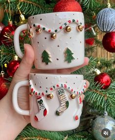 two coffee mugs with christmas decorations on them are held in front of a christmas tree