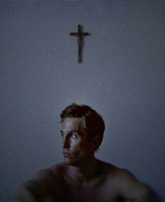 a man sitting in front of a cross on the wall with his shirtless body