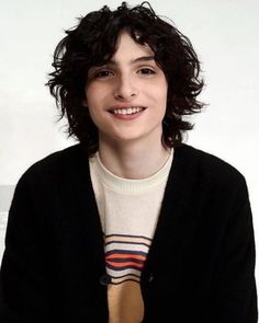 a woman with curly hair wearing a sweater and smiling at the camera while standing in front of a white wall