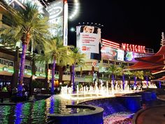 the fountains are lit up at night in front of buildings and palm trees with lights on them