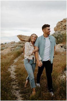 a man and woman holding hands while walking through the grass in front of some rocks