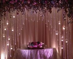 the table is covered with purple flowers and lights in front of a drape curtain