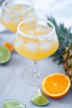 two glasses filled with drinks next to sliced oranges and pineapple on a table