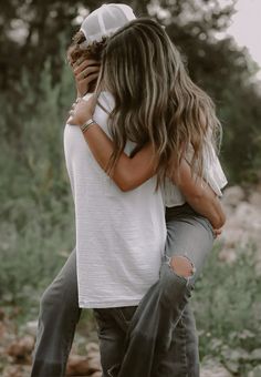 two people hugging each other in front of some rocks and grass with trees behind them