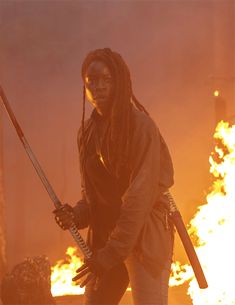 a man with dreadlocks holding two swords in front of a large fire filled background