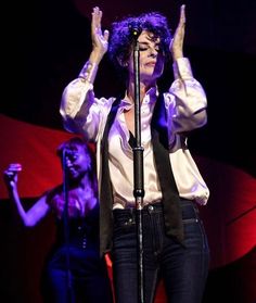 a woman with purple hair holding her hands up in the air while singing into a microphone