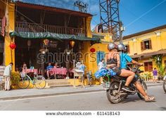 two people riding on the back of a motorcycle in front of a building with yellow walls