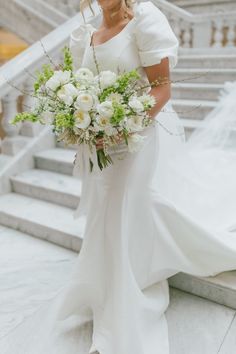 a woman in a white wedding dress holding a bouquet and posing for the camera with stairs behind her