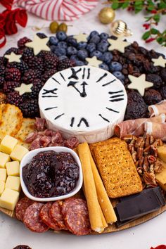 a platter filled with cheese, crackers, fruit and meats next to a clock