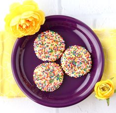 three sprinkled donuts on a purple plate next to a yellow flower and napkin