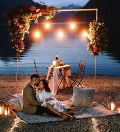 a man and woman sitting on a blanket in front of a lake at night with candles