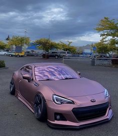 a pink sports car parked in a parking lot under a cloudy sky with trees and bushes