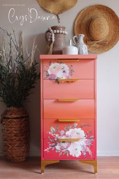 an orange dresser with flowers painted on it next to a potted plant and straw hat