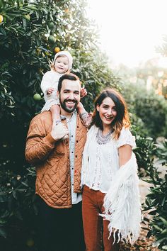 a man and woman standing in front of an orange tree holding a child on their shoulders