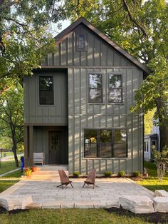 two lawn chairs sitting in front of a gray house with large trees and grass around it