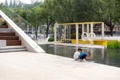 two people sitting on the ground in front of a small pond with water running through it