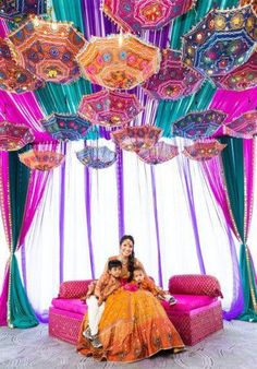 two women sitting on top of a couch under an umbrella covered room with colorful drapes