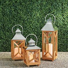 three wooden lanterns sitting next to each other on top of a gravel ground near a hedge