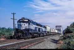 a black and white train traveling down tracks next to trees with blue sky in the background