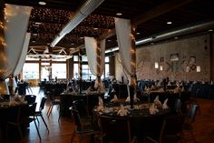 an empty dining room with black tables and white draping on the ceiling is lit by string lights