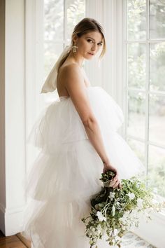 a woman standing in front of a window holding a bouquet of flowers and greenery