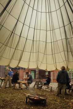 several people sitting around a fire pit under a large tent on the ground with trees in the background