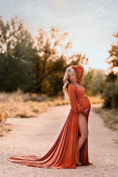 a pregnant woman in an orange dress poses for a photo on a dirt road with trees and bushes behind her