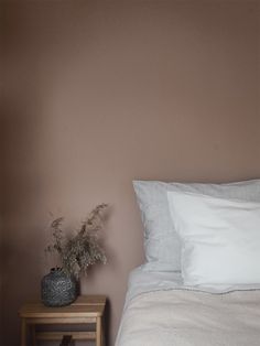 a bed with white sheets and pillows next to a small wooden table holding a plant