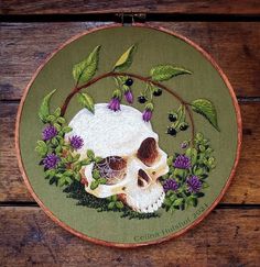 a cross stitch skull with purple flowers and leaves around it on a green circle frame