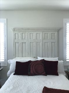 a bed with white headboard and pillows in a bedroom next to two windows that have shutters on them