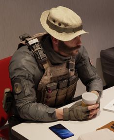 a man sitting at a table in front of a laptop computer wearing a hat and holding a cup