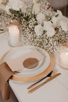 the table is set with white flowers and gold place settings, candles and napkins