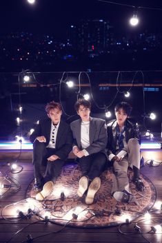 three young men sitting on top of a rug covered in string lights and wearing suits