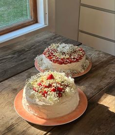 two cakes with strawberries on top sit on plates in front of a windowsill