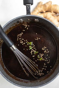 a pan filled with brown liquid next to sliced ginger and an egg whisk