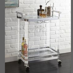 a clear bar cart with liquor bottles on it in front of a white brick wall